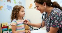 Female pediatrician examining young girl with stethoscope in colorful medical office. Healthcare professional in floral scrubs providing checkup care.