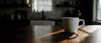 White ceramic mug on wooden table with warm sunlight streaming through window in modern kitchen interior space.
