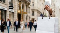 Shopper holding white paper bag on urban street with blurred pedestrians and historic buildings in background, retail and consumer lifestyle.