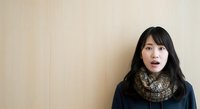 Young East Asian woman with long black hair and knit scarf against neutral beige wall, looking upward thoughtfully. Suitable for lifestyle, education, and corporate projects.
