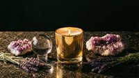 Golden candle glowing warmly surrounded by amethyst crystals, lavender sprigs and clear quartz sphere creating peaceful meditation and wellness atmosphere.
