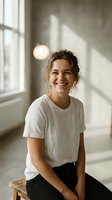 Happy young Caucasian woman laughing in bright modern interior with natural window light wearing casual white t-shirt and black pants sitting on wooden stool.