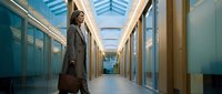 Professional businesswoman walking through modern office corridor with glass walls and natural lighting from skylights above.