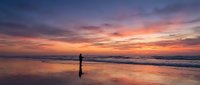 Silhouette of person standing on wet sand beach during golden hour sunset with dramatic colorful sky and ocean waves reflecting warm light on shore.