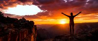 Person with raised arms celebrating sunrise on mountain cliff edge with dramatic orange sky and canyon landscape below for motivational content.