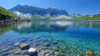 Pristine mountain lake with crystal clear turquoise water reflecting snow-capped peaks, underwater stones visible through transparent alpine water, green meadows on shore.