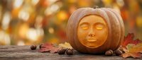 Carved pumpkin with serene face surrounded by autumn leaves and acorns on wooden surface with warm bokeh background for Halloween decoration.