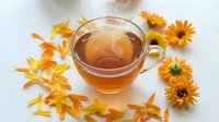 Herbal tea in glass cup surrounded by dried chamomile flowers and petals on white background, wellness and natural remedy beverage.