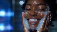 Young African American woman with closed eyes and radiant smile washing face with water splashing, expressing joy during skincare routine in evening light.