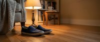 Worn navy blue slip-on shoes on hardwood floor in cozy living room with warm lamp light, sofa, and bookshelf in background.