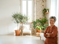 Mature woman with gray hair and Mediterranean ethnicity, wearing a rust-colored cardigan, standing confidently with crossed arms in a bright, plant-filled interior space with natural light and potted greenery for home decor and lifestyle content.