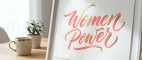Women Power motivational lettering in coral and gold script on white paper displayed in modern interior setting with plant and lamp.