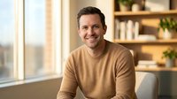 Smiling middle-aged Caucasian man in beige sweater sitting comfortably in modern home office with bookshelf background for lifestyle and business content.