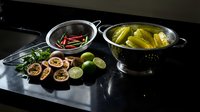 Cooking ingredients arranged on dark surface with fresh limes, red chilies in bowl, boiled potatoes in colander, and sliced citrus for meal preparation.
