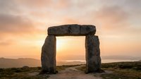 Ancient stone monument with massive rectangular lintel framing golden sunset sky, rolling hills and distant mountains visible through opening, archaeological landmark at dusk.