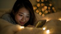 Young Asian woman smiling while using smartphone in cozy bed with fairy lights and Christmas tree bokeh in background, creating warm holiday atmosphere.