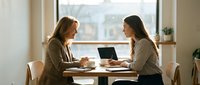 Two professional women having business meeting at wooden table with laptop discussing work collaboration in modern office space.