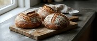 Freshly baked artisan sourdough bread loaves on rustic wooden cutting board in cozy kitchen setting with natural window light for bakery and food photography.