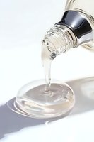 Clear viscous liquid pouring from bottle onto glass surface creates flowing stream with shadow on white background, demonstrating fluid dynamics and transparency.