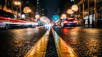 Rainy city street at night with glowing lights, motion blur of passing cars, and wet asphalt reflecting colorful bokeh and yellow road markings.