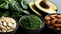 Fresh healthy superfood ingredients with matcha green tea powder, sliced banana, almonds, avocado and spinach leaves arranged on dark background.