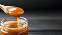 Golden caramel sauce dripping from wooden spoon into glass jar on dark background. Sweet confectionery ingredient for desserts and baking recipes.