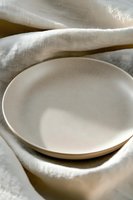 Empty white ceramic plate on soft linen fabric with natural shadows and gentle folds creating minimalist composition for food photography and lifestyle content.