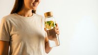Young woman holding glass water bottle with lemon and mint infused water, healthy hydration lifestyle concept on light background.