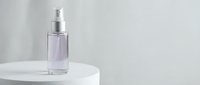 Purple glass spray bottle with silver pump on white pedestal against neutral background for cosmetic and skincare product photography.