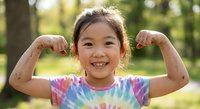 Young Asian girl flexing muscles in tie-dye shirt showing strength and confidence outdoors in sunny park setting for health and fitness concepts.