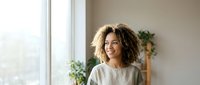 Smiling young African American woman with curly hair standing near window at home, looking away with positive expression.