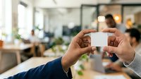 Hands holding blank white business card in modern office environment with colleagues working in background, suitable for corporate branding and professional networking concepts.