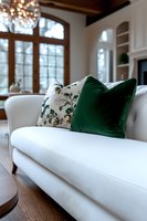 Elegant white upholstered sofa with emerald green velvet and floral pattern throw pillows in luxurious living room featuring arched windows and crystal chandelier.