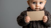 Caucasian baby in knitted sweater holding blank cardboard sign against gray background. Space for text on card and right side of photo.