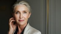 Elegant mature woman with silver hair in light beige outfit against neutral background, touching face gently with thoughtful expression.