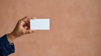 Hand holding blank white business card against neutral beige background, suitable for branding, corporate identity, and marketing mockup designs.