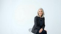 Mature woman with gray hair and fair complexion wearing dark blazer, sitting against white background, smiling at camera for corporate headshot or business portrait use.