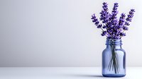 Fresh lavender stems arranged in blue glass mason jar against light neutral background, minimalist floral composition with copy space for wellness designs.