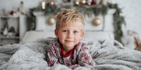 Smiling young Caucasian boy in pajamas lying on cozy gray bedding with festive Christmas decorations in background.