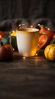 Ceramic mug with warm beverage surrounded by autumn gourds, small pumpkins, and glowing string lights on rustic wooden surface for fall season marketing.