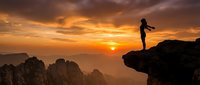 Woman hiker silhouette standing on mountain cliff edge at golden sunrise with arms outstretched celebrating achievement over dramatic landscape vista.