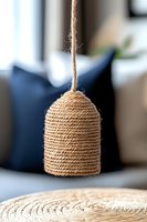 Handcrafted pendant lamp shade made from natural jute rope wrapped in cylindrical shape, hanging against blurred modern interior background with navy cushions.