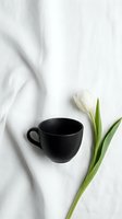 Matte black ceramic coffee cup beside a white tulip on soft linen fabric, minimal flat lay composition for lifestyle and blog use.
