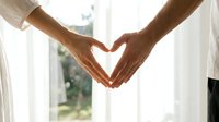 Two people forming heart shape with hands together in bright window light creating romantic love symbol for relationships and connection.