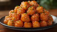 Glazed Asian meatballs stacked on black ceramic plate, garnished with sesame seeds and fresh chopped herbs, served with sweet and savory sauce.