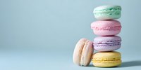 Stack of colorful French macarons in pastel shades of mint green, pink, lavender, and yellow against a soft blue background.