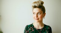 Blonde woman with upswept hairstyle wearing dark floral top and necklace, looking away with confident expression against neutral background.