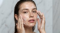 Young woman applying white facial cleanser foam with both hands during morning skincare routine against marble background for beauty and wellness concepts.