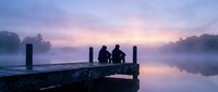 Two people sitting on wooden dock at misty lake during purple sunrise reflecting peaceful morning atmosphere for relaxation and friendship concepts.