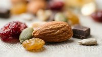 Mixed nuts, dried fruits and dark chocolate pieces arranged on rustic linen fabric. Healthy snacking ingredients for trail mix, granola or energy bars preparation.